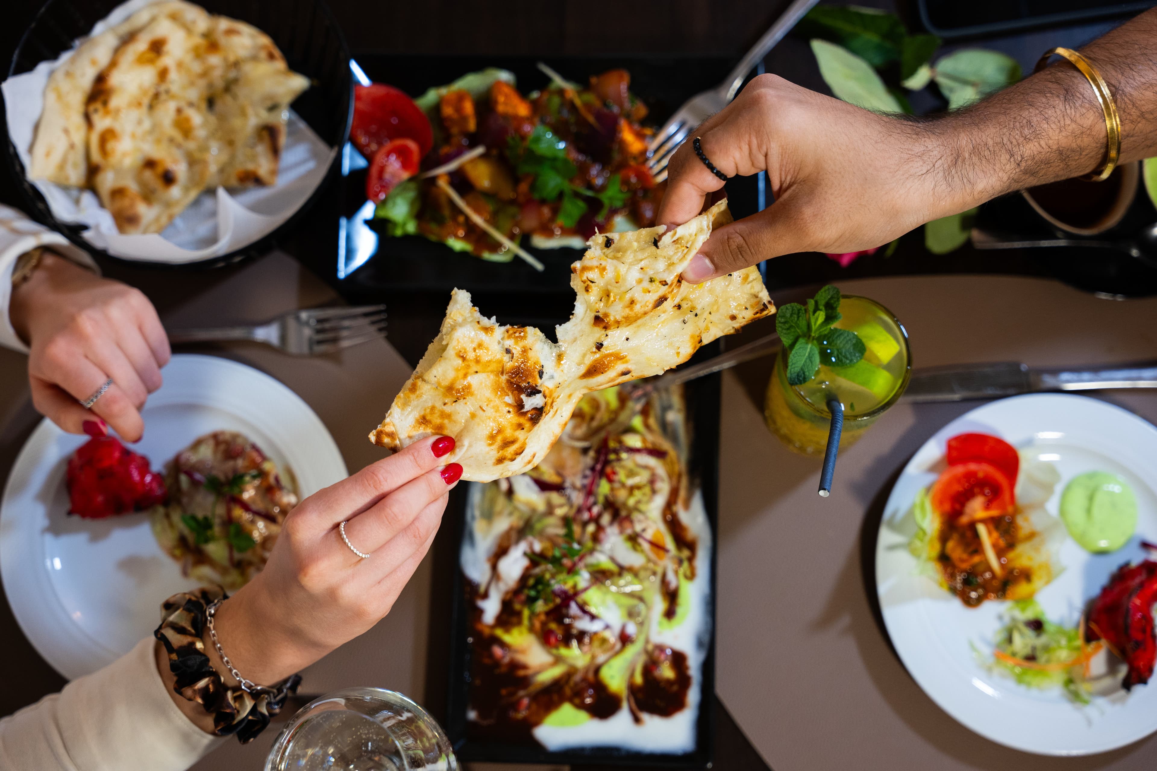 Hände teilen frisch gebackenes Naan-Brot über einer Auswahl indischer Gerichte